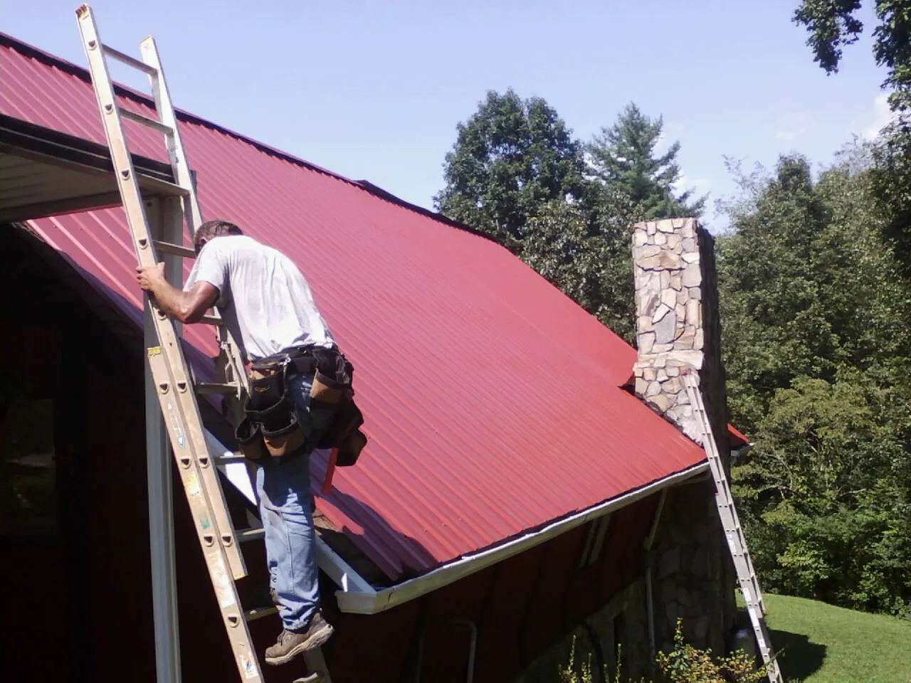 Roofer on a ladder beside a red metal roof and stone chimney — Soffit & Fascia Repair by Roofing Brothers in Chaplin